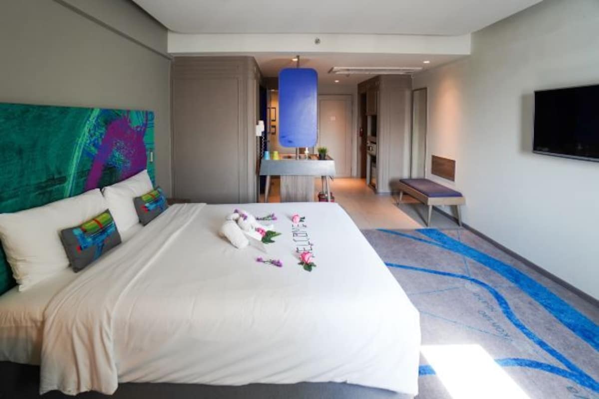 Una habitación de hotel moderna con una cama grande, un cuadro abstracto colorido en la pared, un televisor de pantalla plana y un baño visible a través de una puerta abierta.