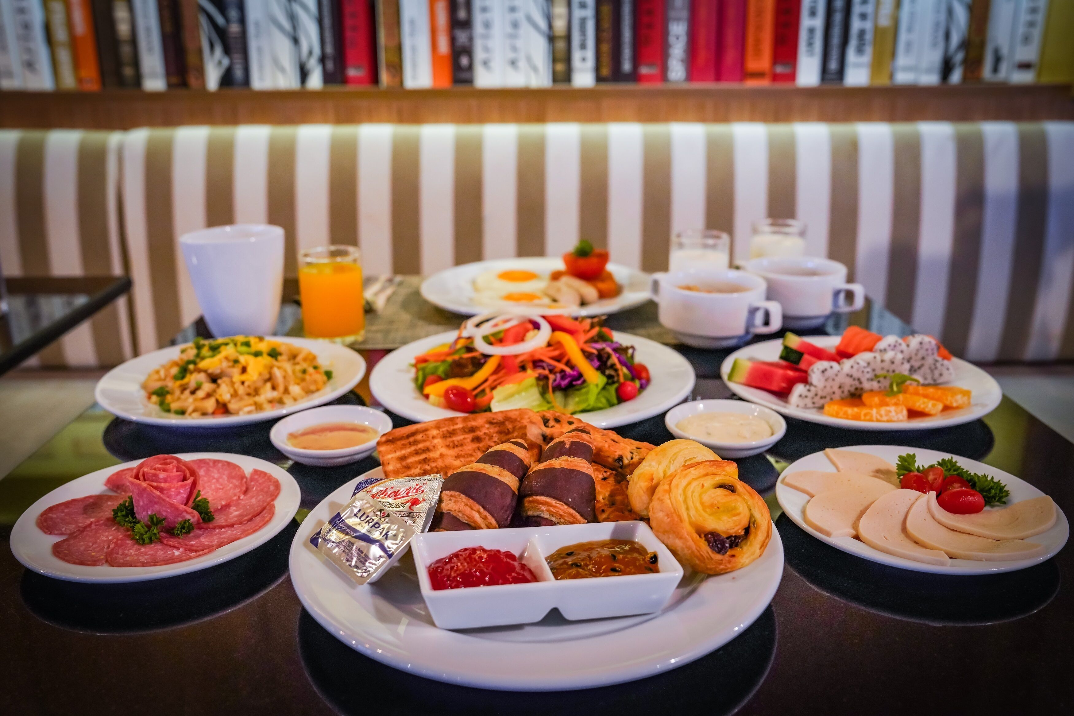 A breakfast buffet with various dishes, including meats, pastries, and fruits, on a table with striped cushions in the background.