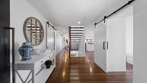 A modern hallway with a wooden floor, white walls, and a central staircase. - Victoria's - Hamptons Charm, Pool, Pizza Oven, Spectacular Ocean Views (Emerald Beach)
