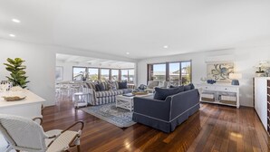A spacious living room with a large sofa, a coffee table, and a dining area with chairs. - Victoria's - Hamptons Charm, Pool, Pizza Oven, Spectacular Ocean Views (Emerald Beach)