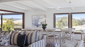 A modern living room with a sofa, chairs, a dining table, and a large window with a view of the outdoors. - Victoria's - Hamptons Charm, Pool, Pizza Oven, Spectacular Ocean Views (Emerald Beach)