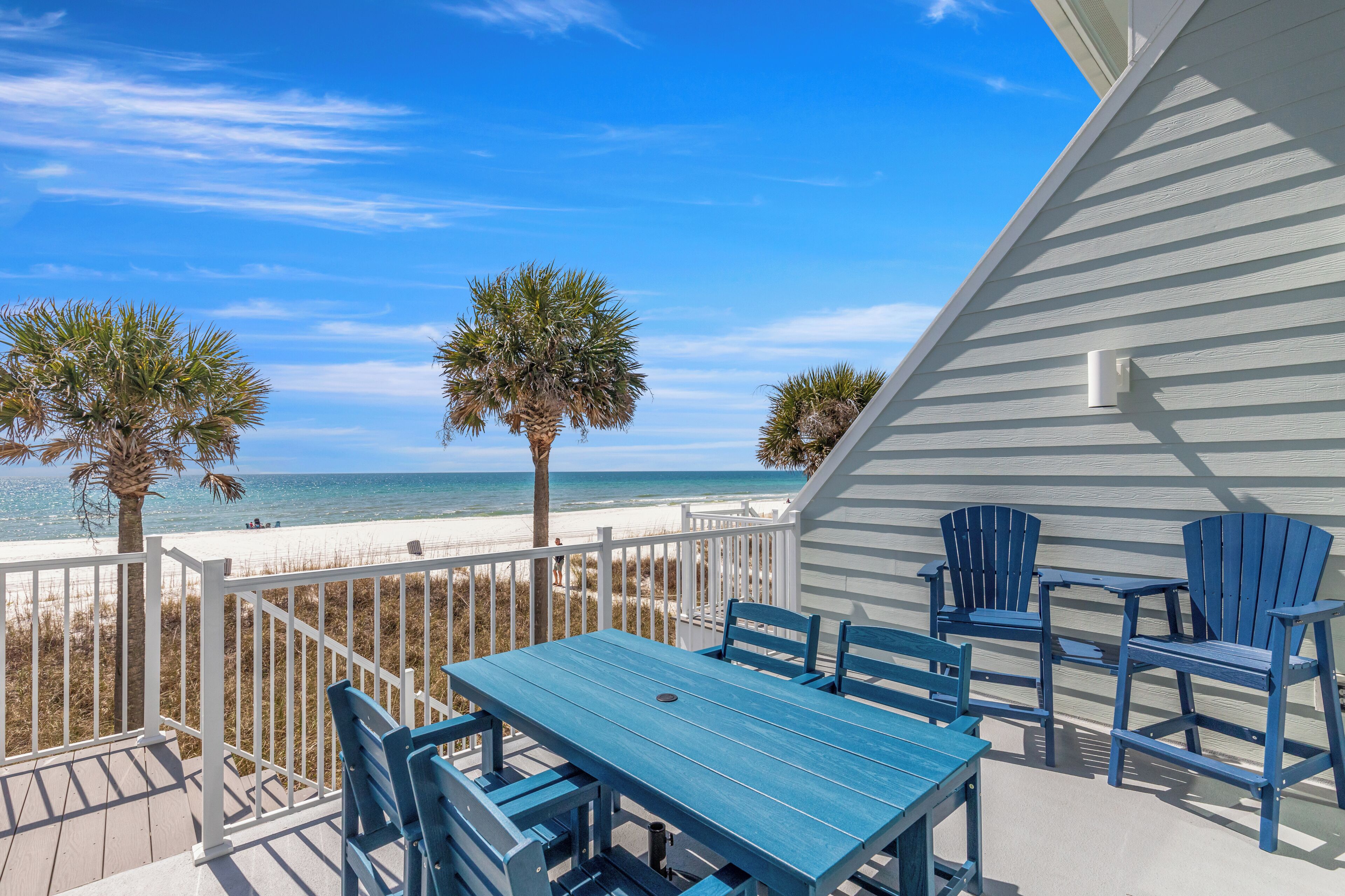 A deck with a table and chairs, overlooking a beach with palm trees and a white fence.