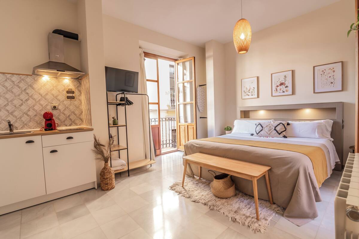 A modern bedroom with a large bed, bedside table, and a small desk. There is a kitchen area with a stove, sink, and cabinets. The room has a large window and a door leading to a balcony.