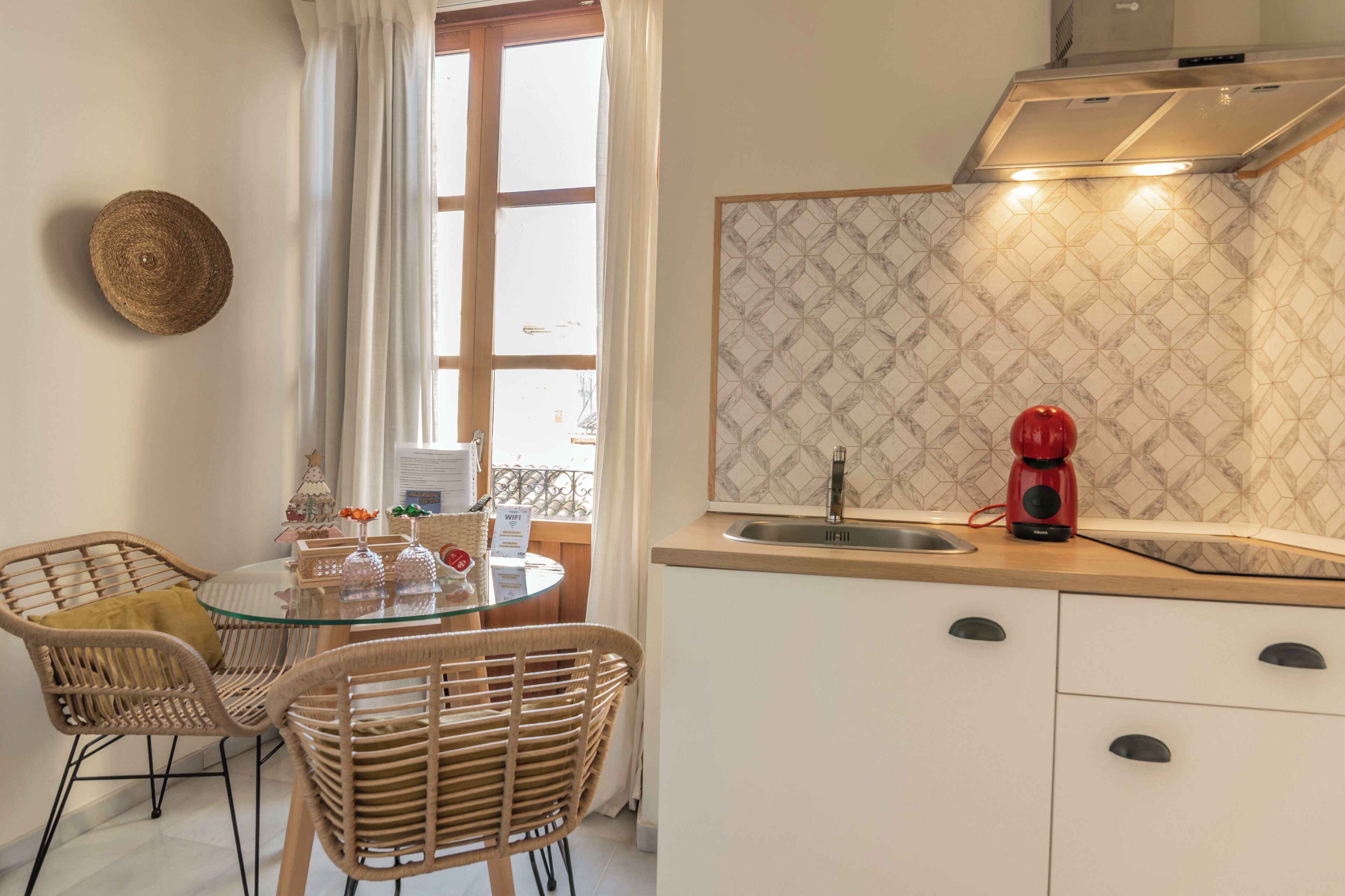 A kitchen with a round glass table, wicker chairs, a red coffee maker, and a window with white curtains.