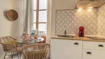 A kitchen with a round glass table, wicker chairs, a red coffee maker, and a window with white curtains.