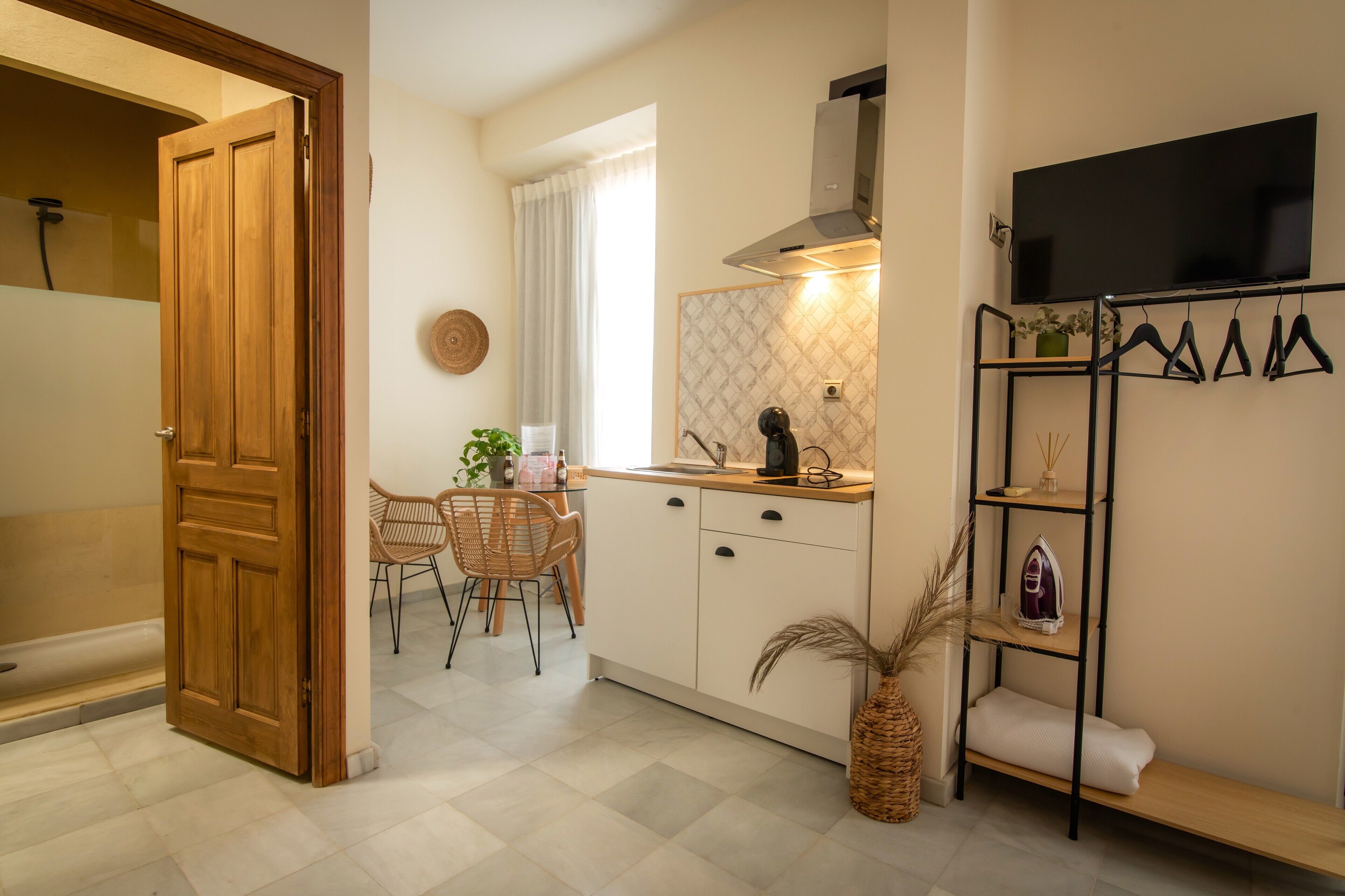 A compact kitchen with a dining area, a wooden door, a TV mounted on the wall, and a shelf with hanging items.