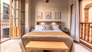 A bedroom with a large bed, a wooden bench, a small table, a hanging light fixture, and a balcony with a wrought iron railing. - APARTAMENTOS VITA CENTER  (Granada)