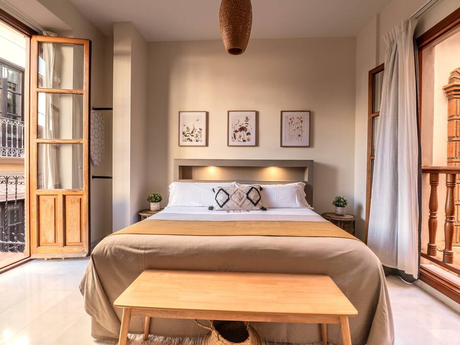 Un dormitorio con una cama grande, un banco de madera, una mesita, una lámpara colgante y un balcón con barandilla de hierro forjado.
