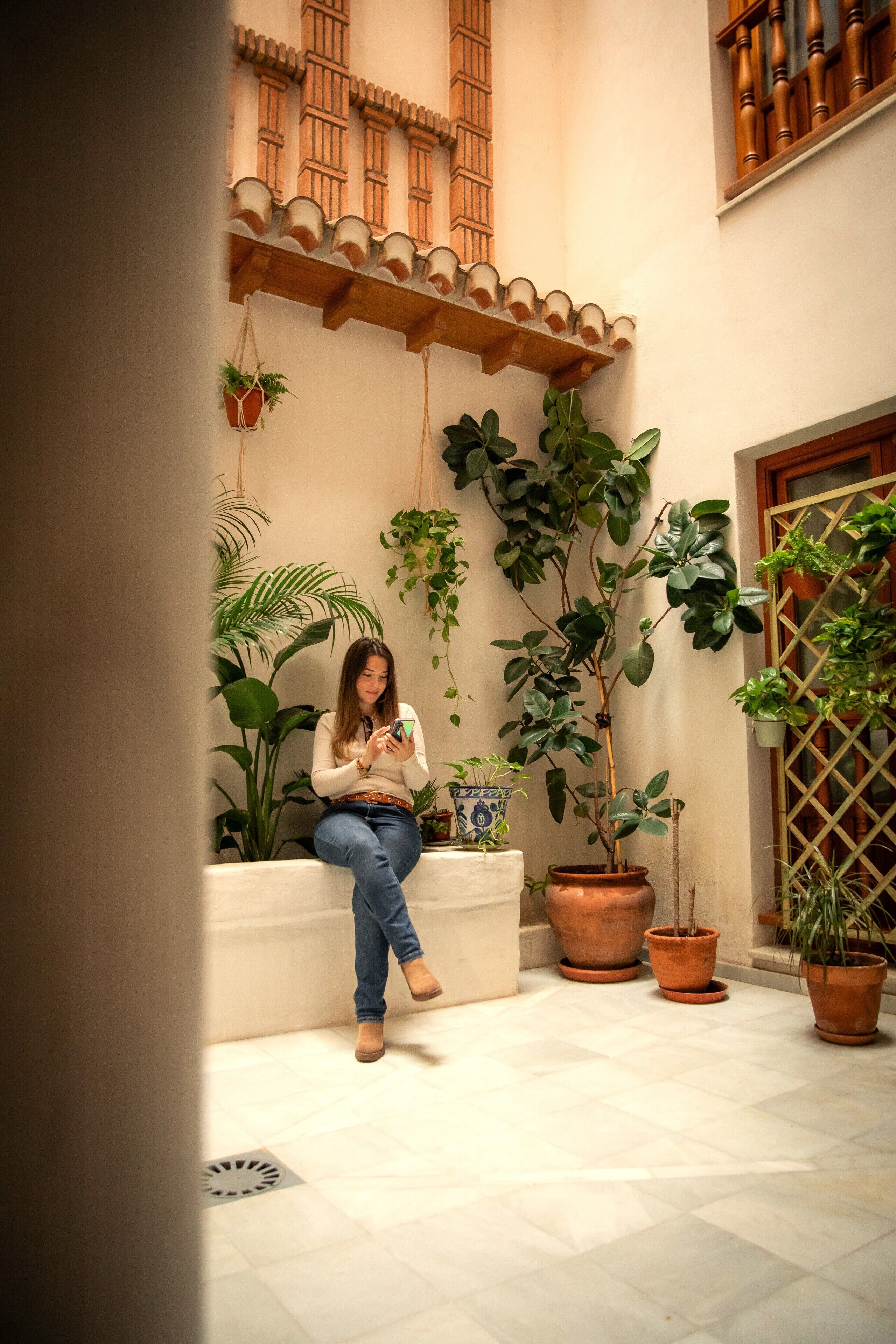 Una mujer sentada en un banco en un patio con plantas en macetas y suelo de baldosas.