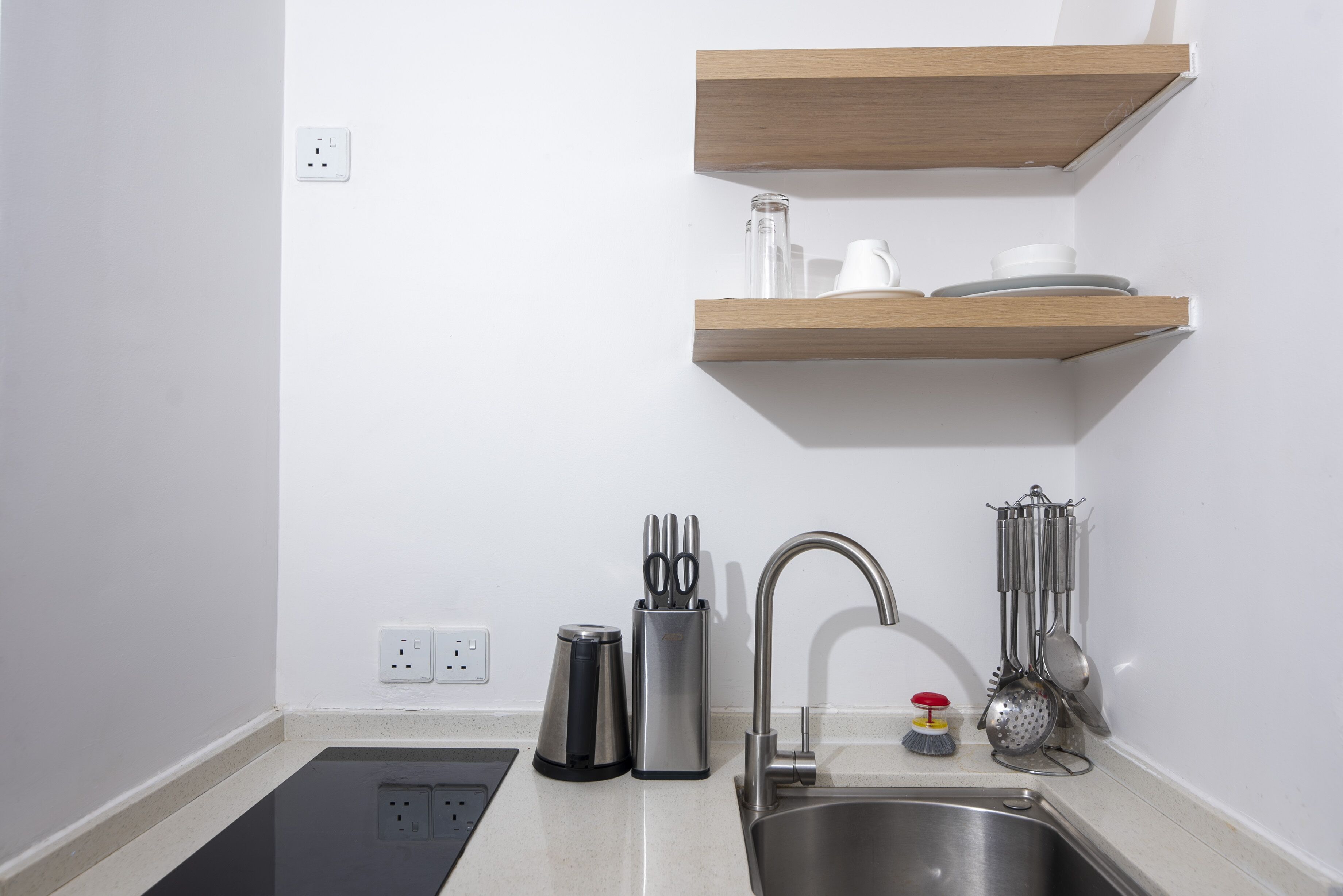 A modern kitchen with a stainless steel sink, wooden shelves above, and a countertop with a coffee maker, knife block, and utensils.