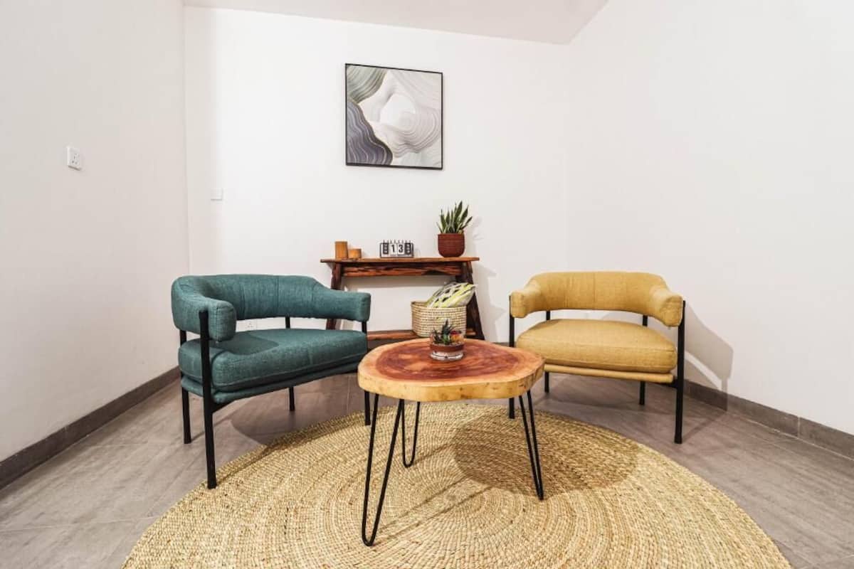 Een kamer met een ronde houten tafel, twee stoelen, een klein plankje met een plant en een abstract schilderij aan de muur.