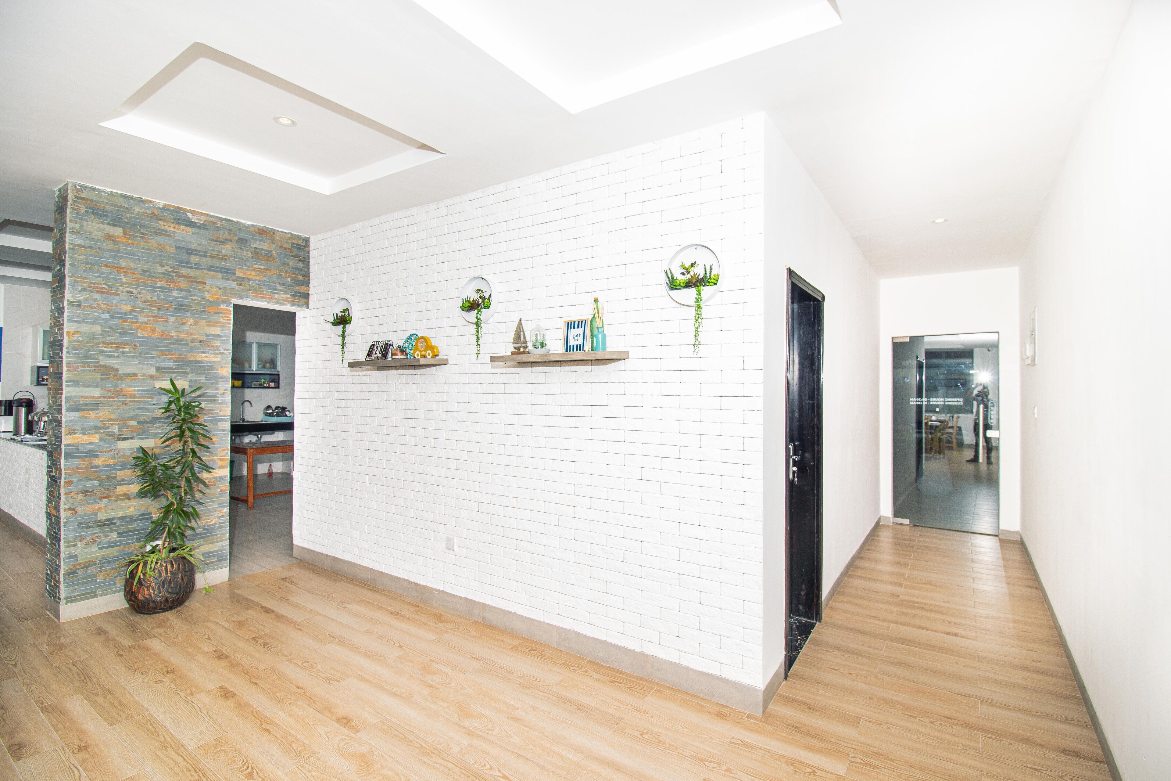 A modern interior with a white brick wall, wooden flooring, and a kitchen area visible through an open doorway.