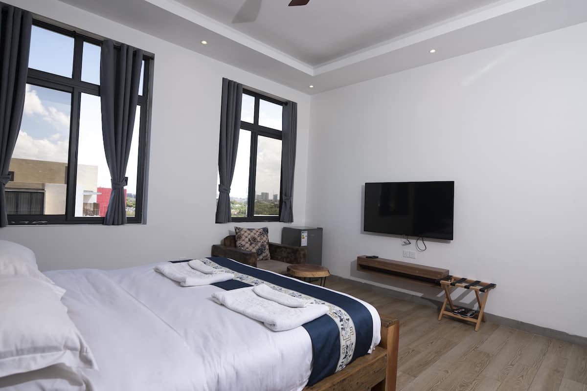 Een moderne hotelkamer met een groot bed, een flatscreen televisie, een zithoek met een bankje, en grote ramen met uitzicht over de stad.