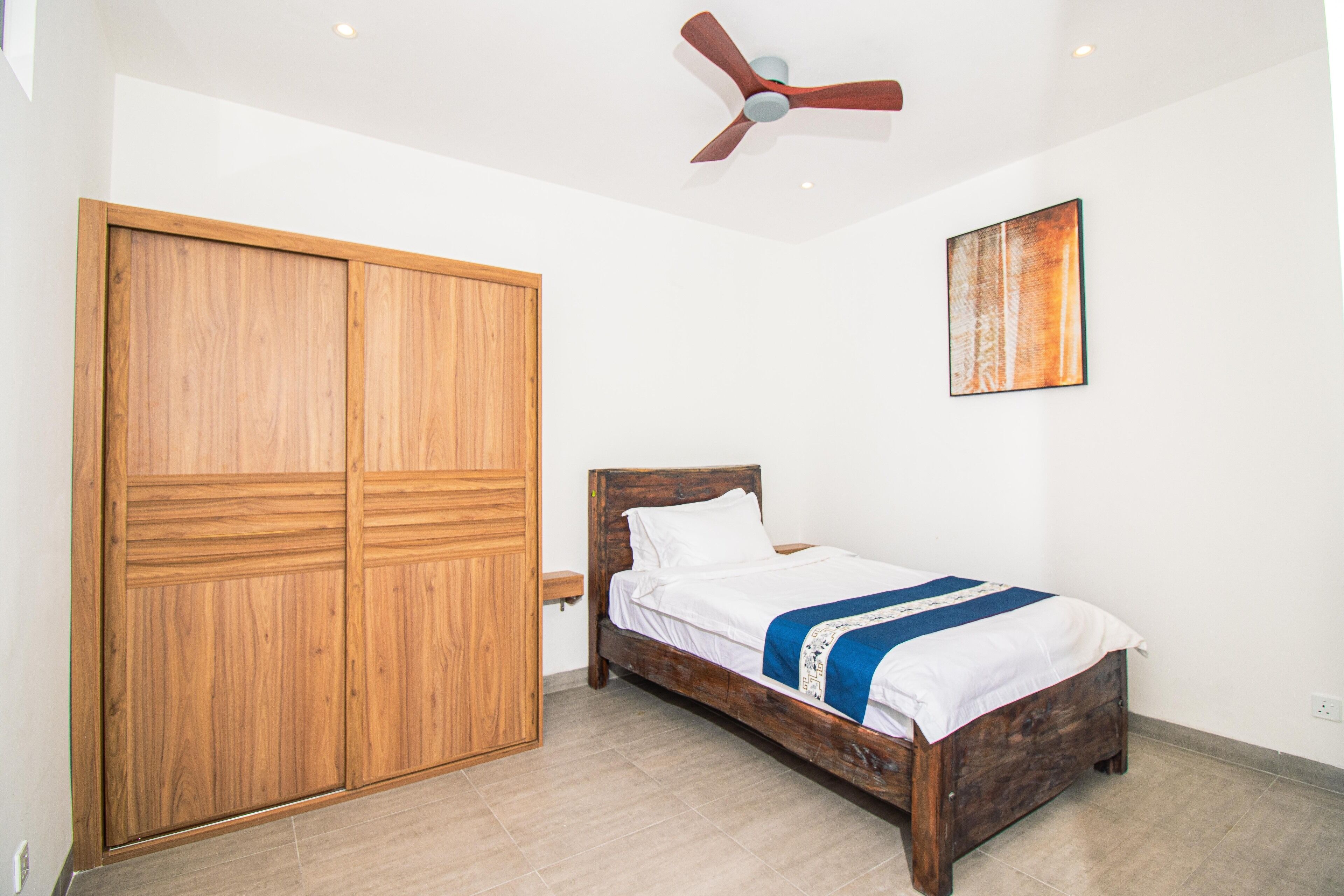 A bedroom with a wooden wardrobe, a bed with a blue and white bedspread, a ceiling fan, and a painting on the wall.