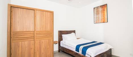 Un dormitorio con un armario de madera, una cama con colcha azul y blanca, un ventilador de techo y un cuadro en la pared.