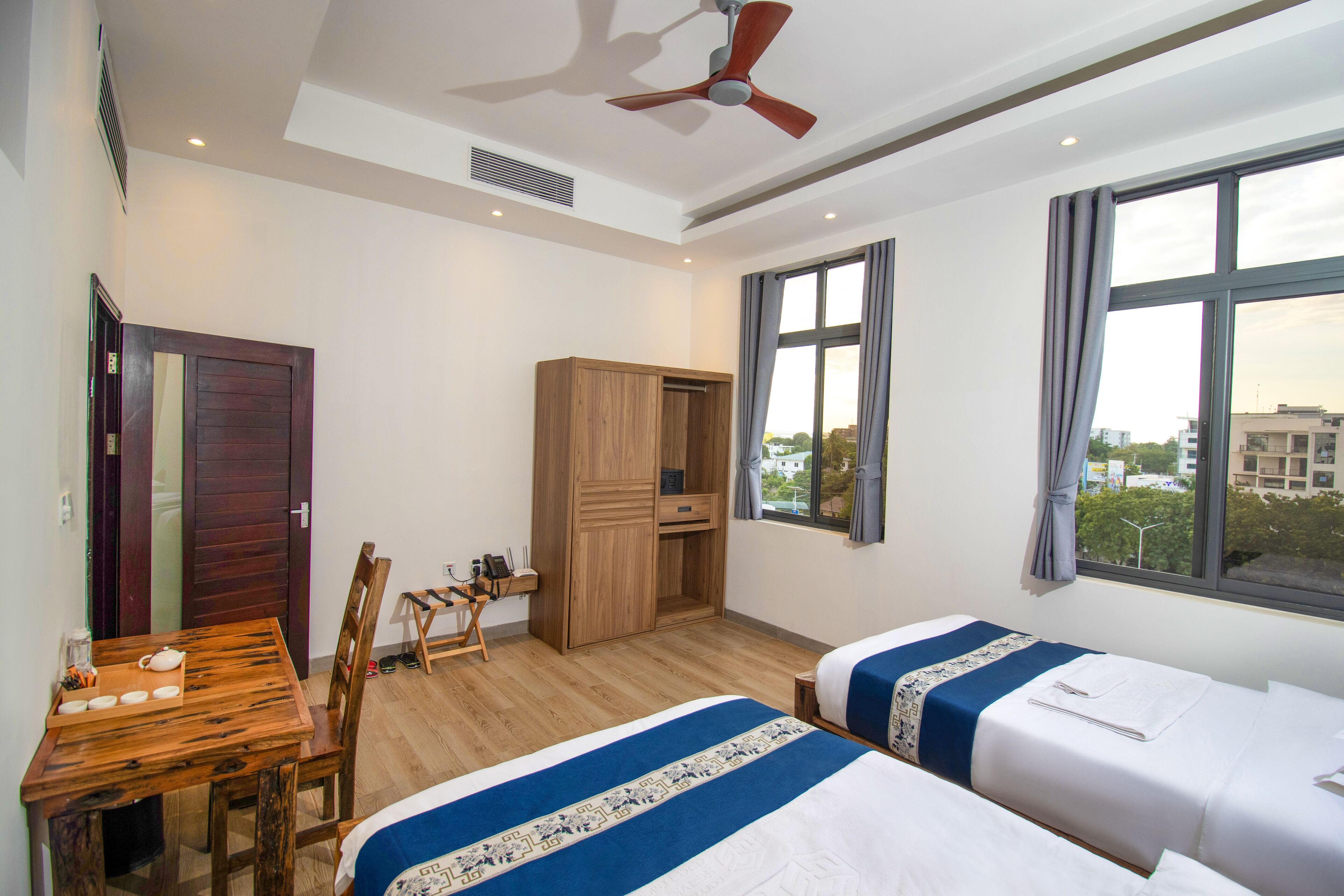 A hotel room with two beds, a wooden desk, a wardrobe, and a view of the city through the window.