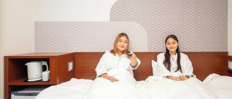 Two people sitting on a bed with a wooden headboard, a coffee maker, and a small shelf with a kettle and mug.