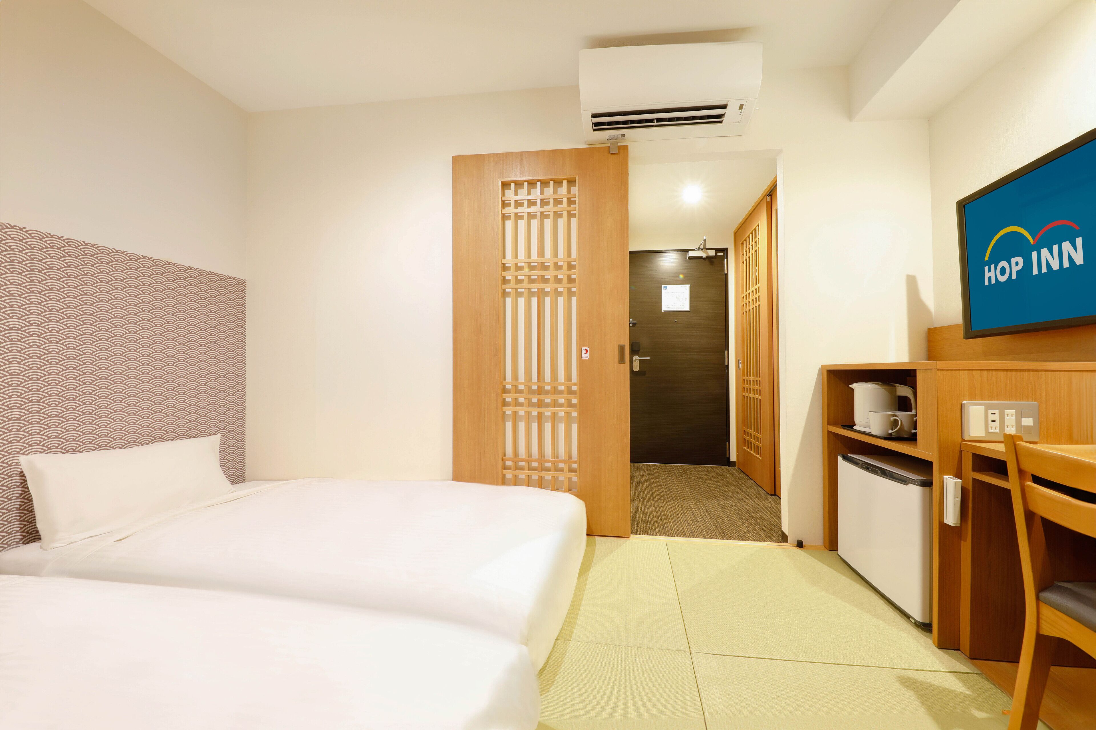 A hotel room with a bed, a desk with a chair, a small refrigerator, and a television displaying the Hop Inn logo.