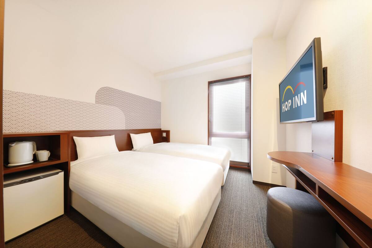 A hotel room with two beds, a TV, a desk, a mini-fridge, and a window with blinds.