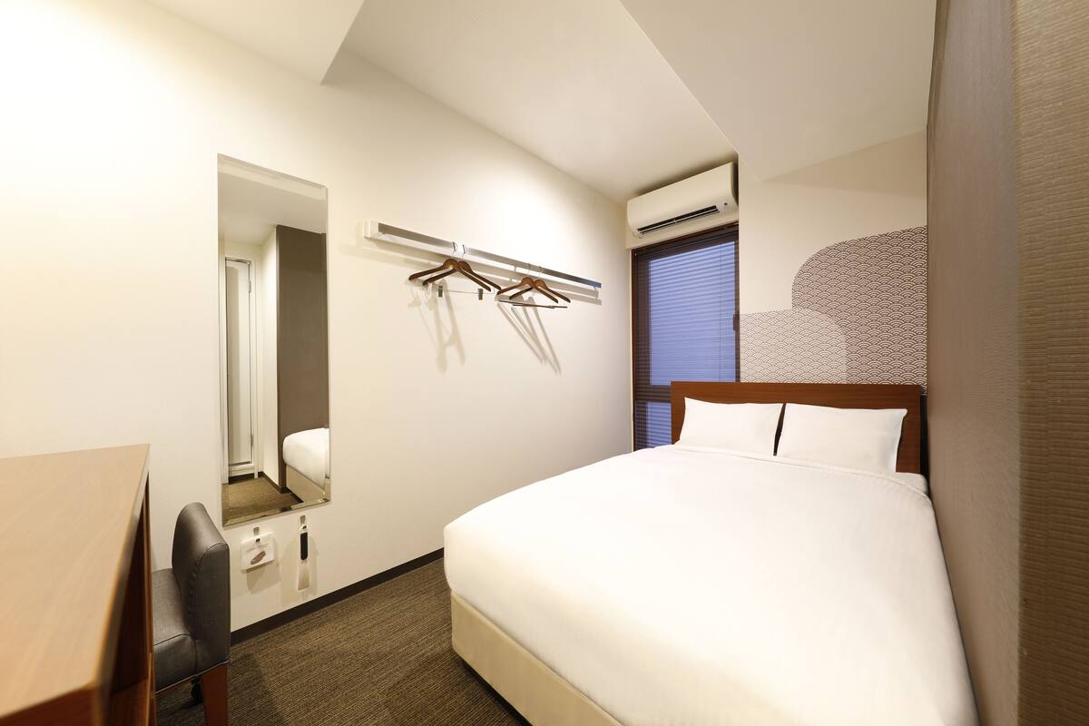 A hotel room with a bed, a desk, a chair, a mirror, a wardrobe, and an air conditioning unit.