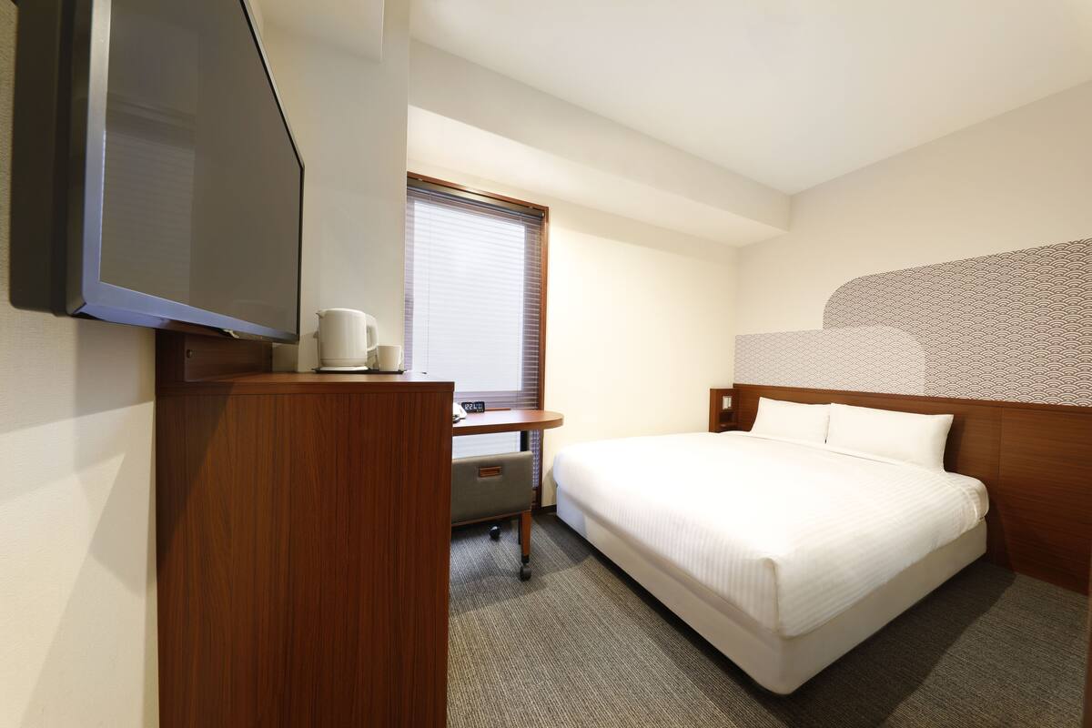 A hotel room with a bed, a desk, a television, and a window with blinds.