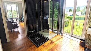 A modern bathroom with a glass shower enclosure, wooden flooring, and a view of a green garden through large windows.