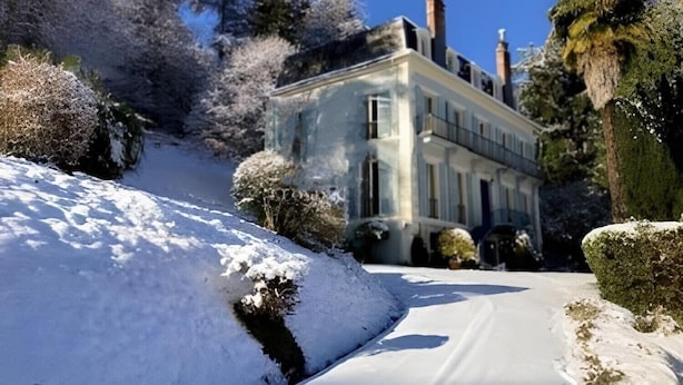 A snow-covered landscape with a large building, possibly a hotel, surrounded by trees and bushes.