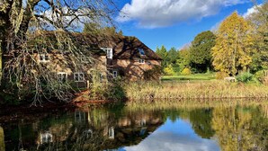 A house by a calm lake with a clear reflection, surrounded by trees and a grassy area.