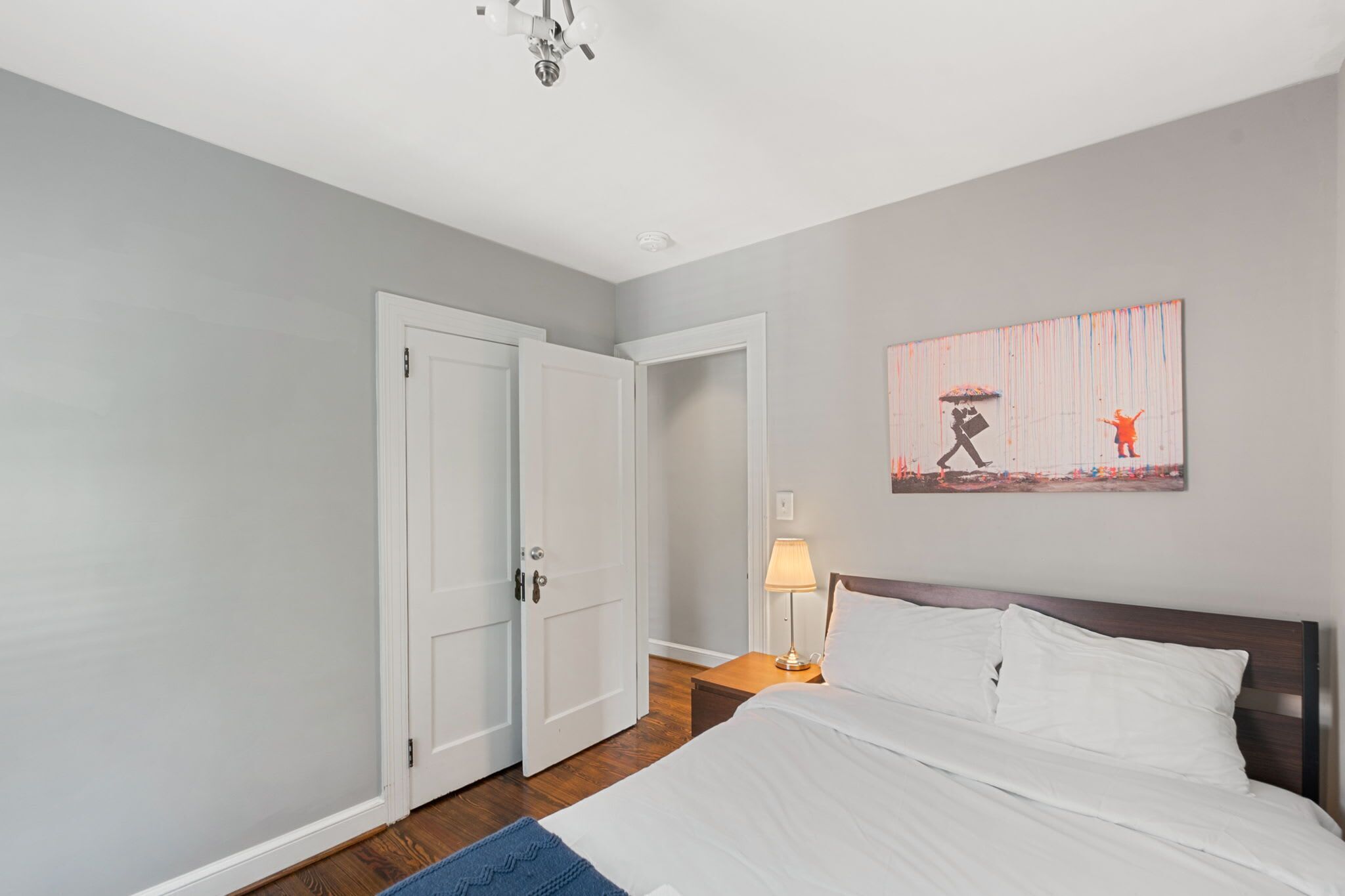 A bedroom with a bed, a nightstand with a lamp, a door, and a wall-mounted artwork.