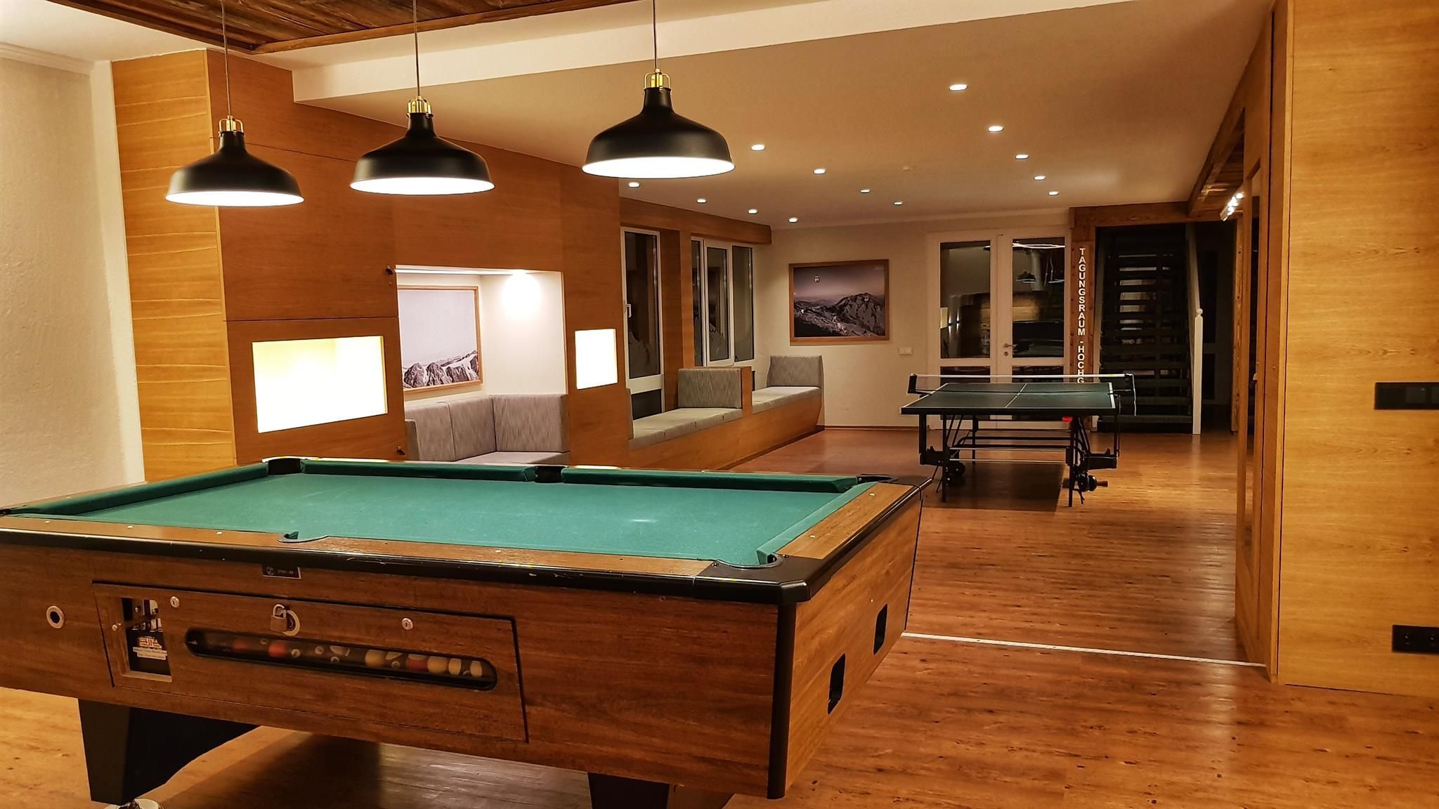 A pool table in a room with wooden flooring, recessed lighting, and a seating area with a sofa and cushions.