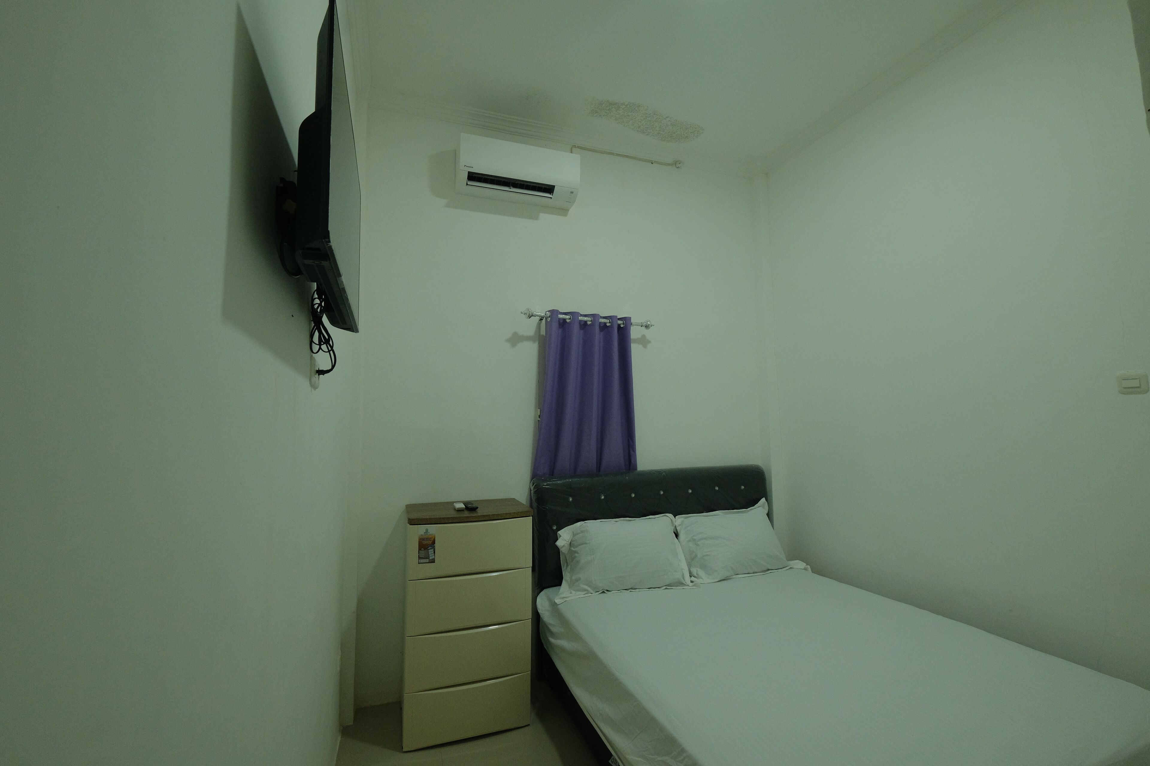 A small, single-bed room with a bed, a nightstand, a TV mounted on the wall, and an air conditioning unit.