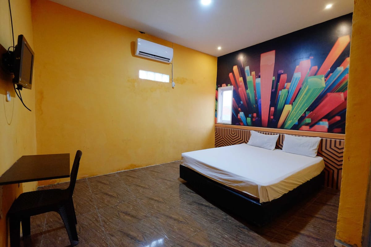 A hotel room with a bed, a desk, a chair, an air conditioner, and a colorful abstract wall mural.