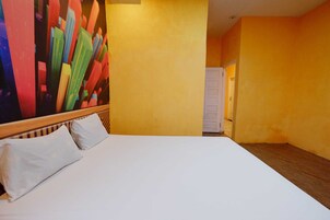 Quarto de hotel com uma cama grande, roupa de cama branca e um quadro abstrato colorido na parede.