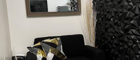 A modern living room with a black sofa, a fluffy grey rug, a geometric black wall panel, and a mirror reflecting a clock and a geometric wall panel.