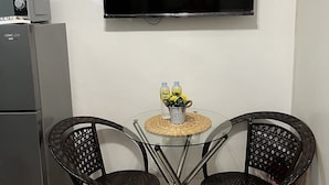 A compact living space with a flat-screen TV mounted on the wall, a small refrigerator, and two wicker chairs around a glass table with a woven placemat. - Lovely 1BR Condo SM Southmall 100mbps Netflix Disney+ (Las Piñas)