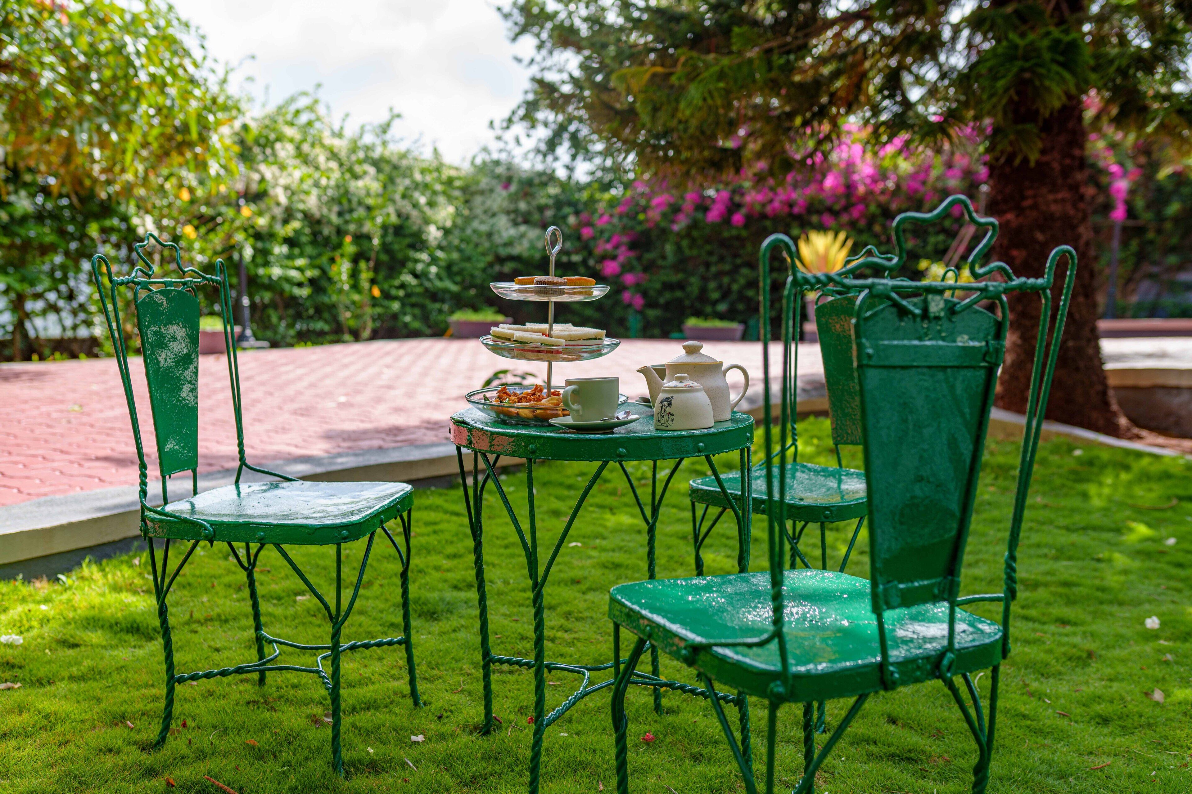 A garden setting with a table and chairs, a tiered tray with food, a teapot, and a cup.