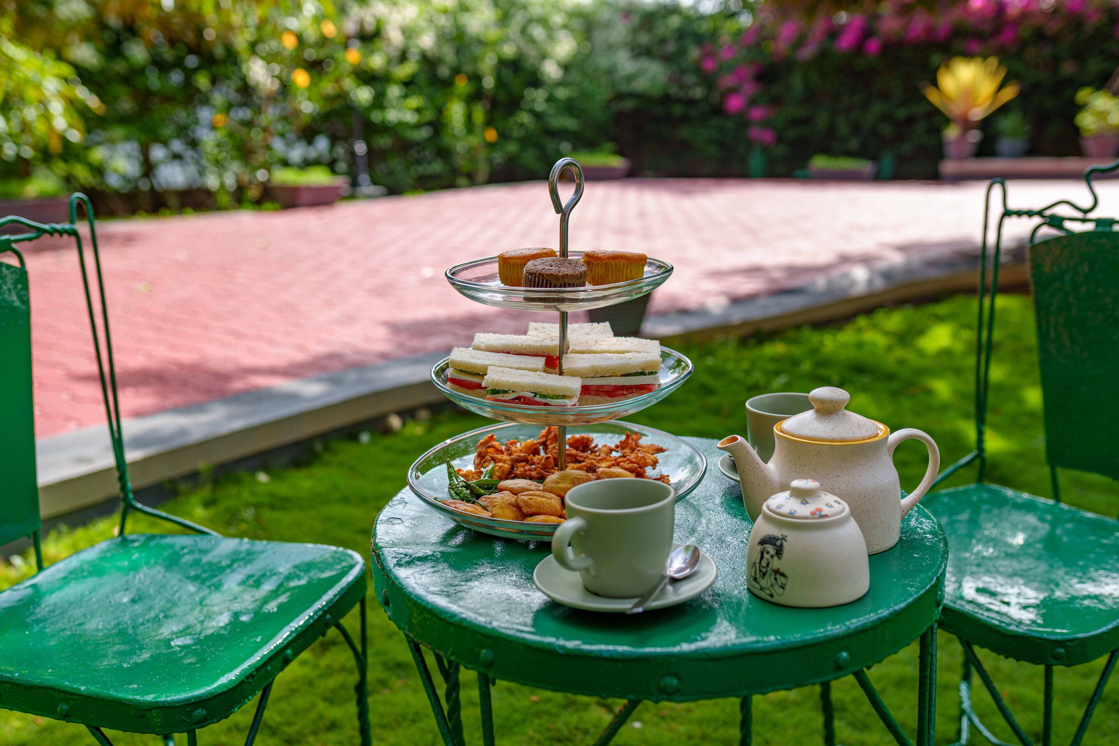 A three-tiered dessert stand with sandwiches and pastries, a teapot, cups, and saucers on a green table in a garden setting.