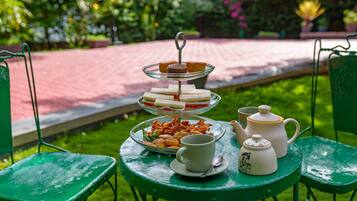 A three-tiered dessert stand with sandwiches and pastries, a teapot, cups, and saucers on a green table in a garden setting.