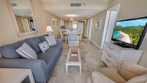 A living room with a grey sofa, a beige armchair, a coffee table, a TV on a stand, and a view of a beach through a window. - Stunning Views 2BR Oceanfront, Beach Cove & Pools (North Myrtle Beach)