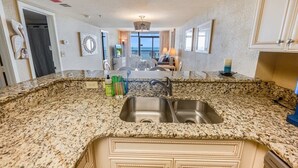 A kitchen with granite countertops, a double sink, and a view to the living area. - Stunning Views 2BR Oceanfront, Beach Cove & Pools (North Myrtle Beach)