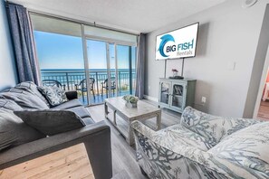 Una sala de estar moderna con un sofá, un sillón estampado y una mesa de centro. Un televisor montado en la pared muestra 'BIG FISH Rentals'.