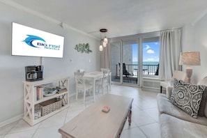 A modern living room with a flat-screen TV displaying a logo, a dining area with white furniture, and a balcony with a view of the sea.