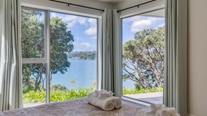 A room with a large window overlooking a lake, a bed with rolled towels, and a view of trees and hills. - Absolute Beachfront - Oneroa Bay (Auckland)