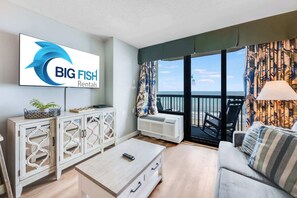 Una sala de estar moderna con un televisor grande mostrando el logotipo de 'BIG FISH Rentals', un balcón con vistas y un área de descanso acogedora.
