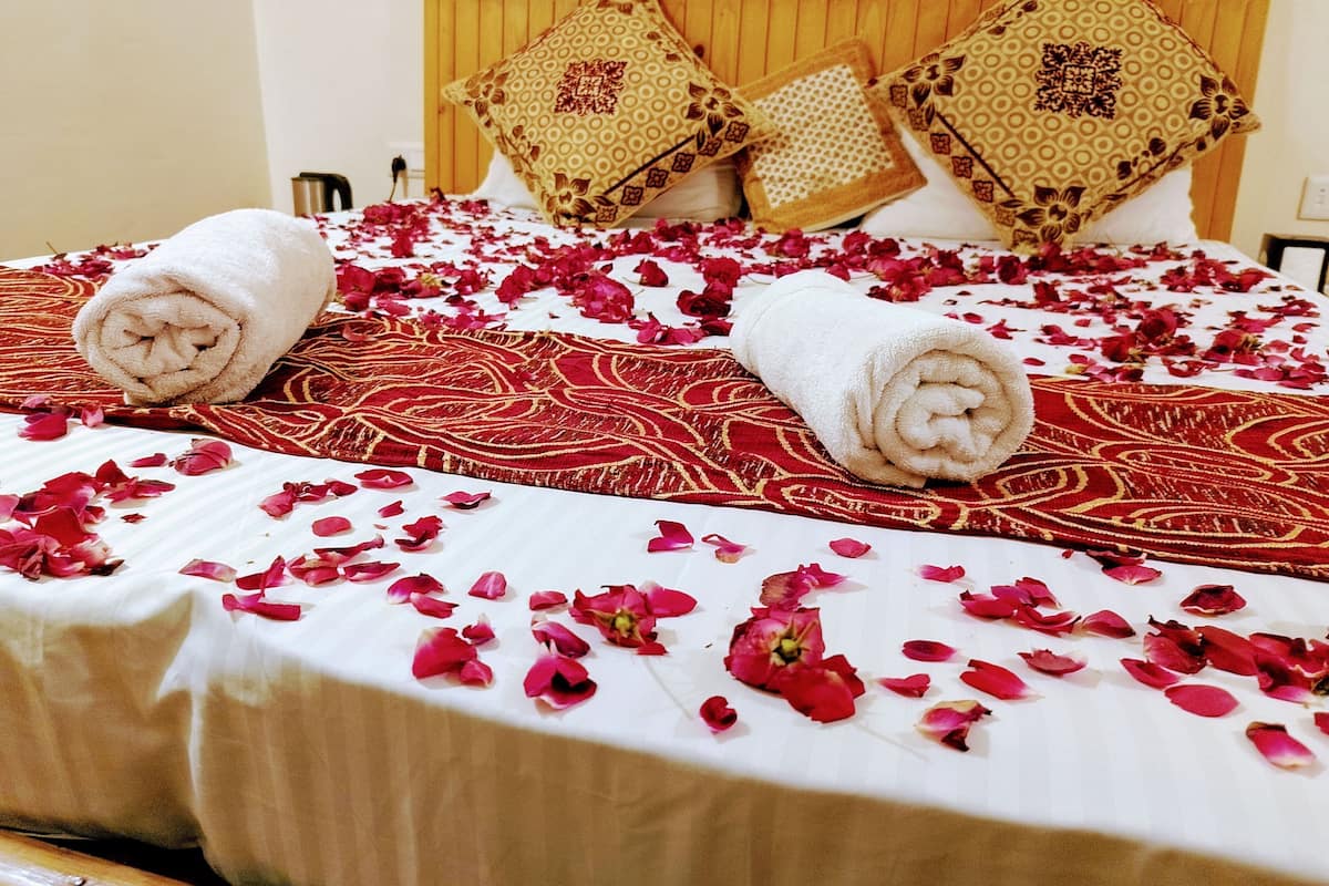Una cama bien hecha con toallas enrolladas y pétalos de rosa esparcidos, un cabecero de madera y almohadas estampadas.