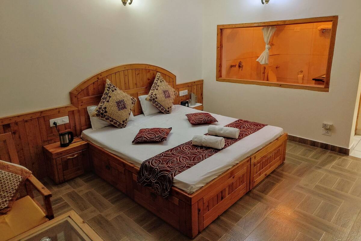 Una cama de madera con almohadas estampadas, una mesita de noche con hervidor y una ventana con cortinas.