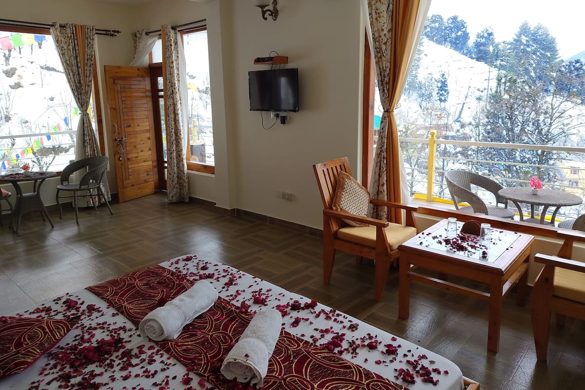 Una habitación con cama, televisión, balcón con sillas y una mesa decorada con pétalos de rosa.