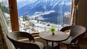 Een kamer met uitzicht op besneeuwde bergen en een stadje. Er staat een rond tafel met twee rieten stoelen en een vaas met bloemen.