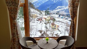 Een kamer met een groot raam met uitzicht op een besneeuwd berglandschap. Er staat een ronde tafel met twee rieten stoelen en een kleine plant in pot op de tafel.