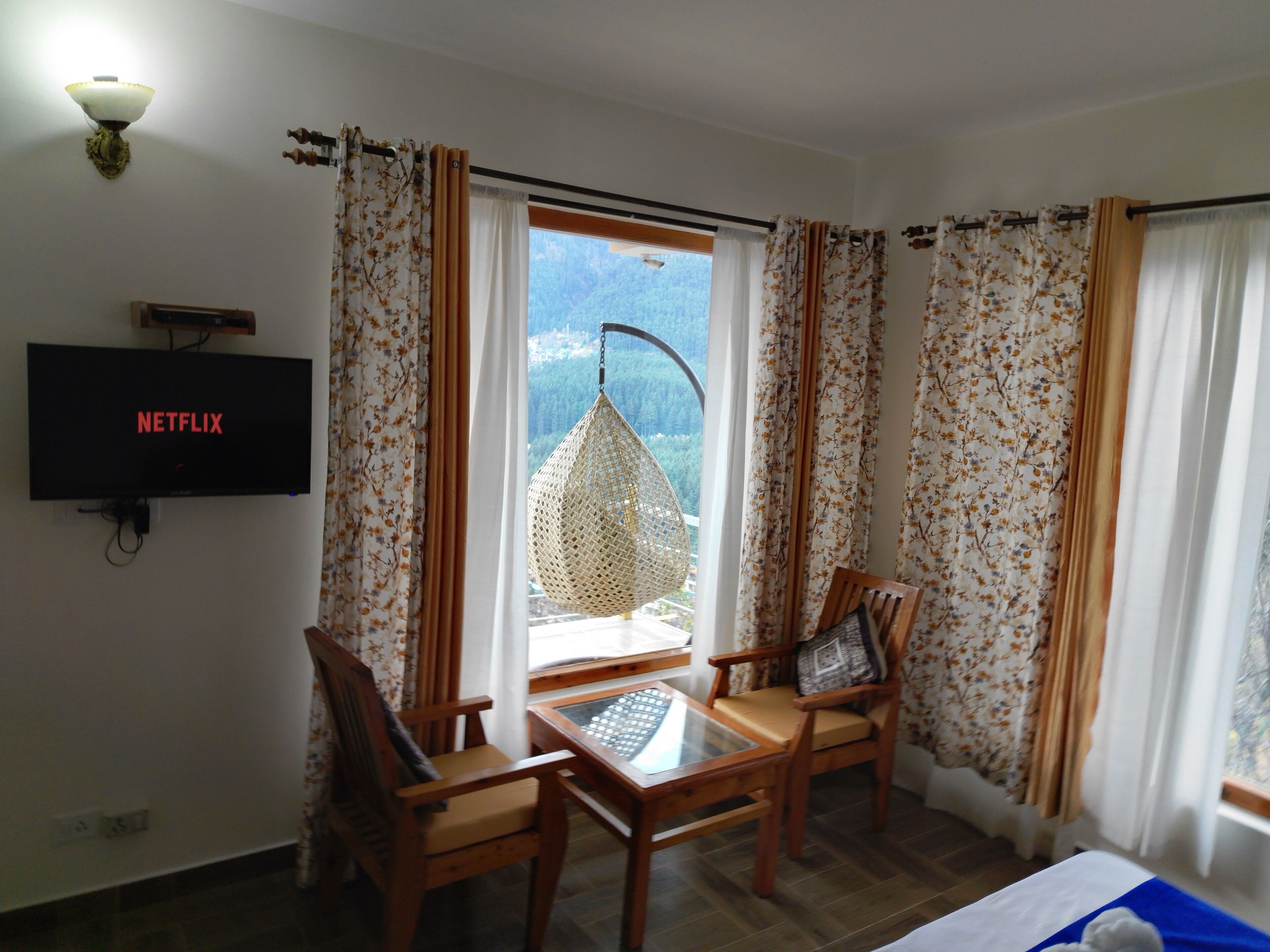 A room with a bed, a TV displaying 'NETFLIX', a table with chairs, and a view of a mountainous landscape through the window.
